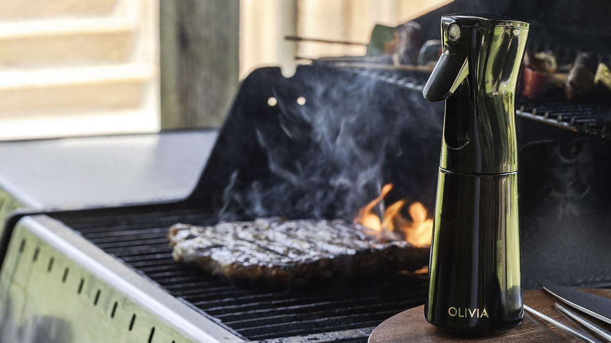 Black oil sprayer for cooking on a wooden board beside a gas grill with a steak searing