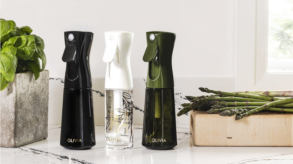 Black, clear and olive green glass oil spray bottle lineup on a white marble counter between a pot of basil and fresh asparagus