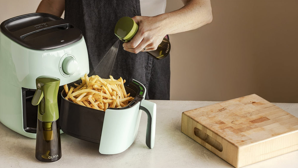Hand using a green olive oil sprayer to mist fries in an open air fryer basket