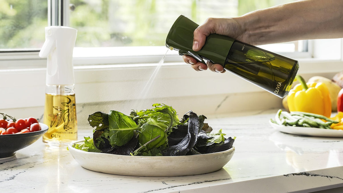 Hand using a green olive oil spray bottle to mist a plate of leafy salad on a bright kitchen counter