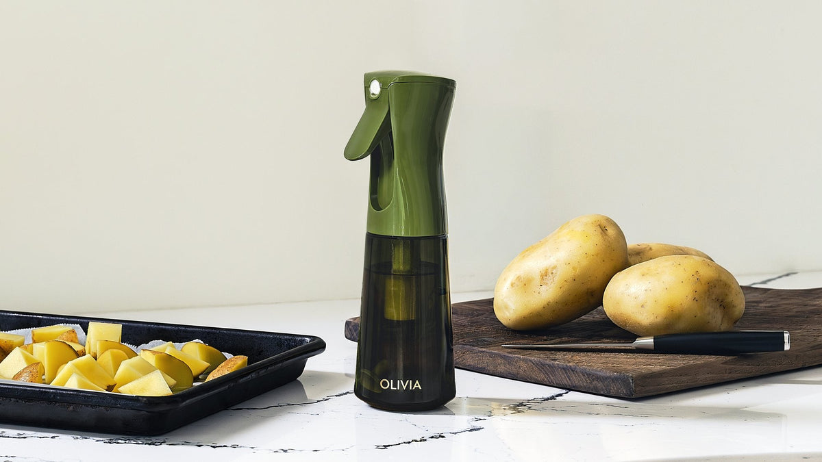 Green olive oil sprayer on a marble counter between a baking tray of cubed raw potatoes and whole potatoes on a dark wooden board