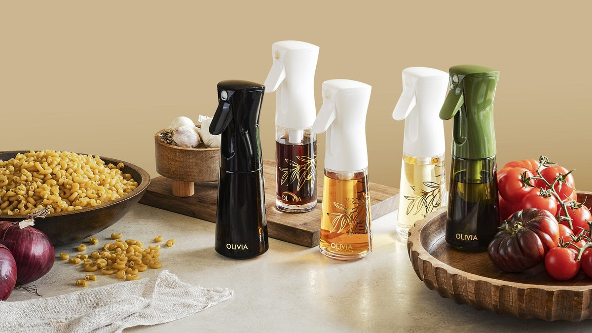 Five glass olive oil spray bottles in black, white and green filled with different oils, beside tomatoes, red onions and a bowl of pasta
