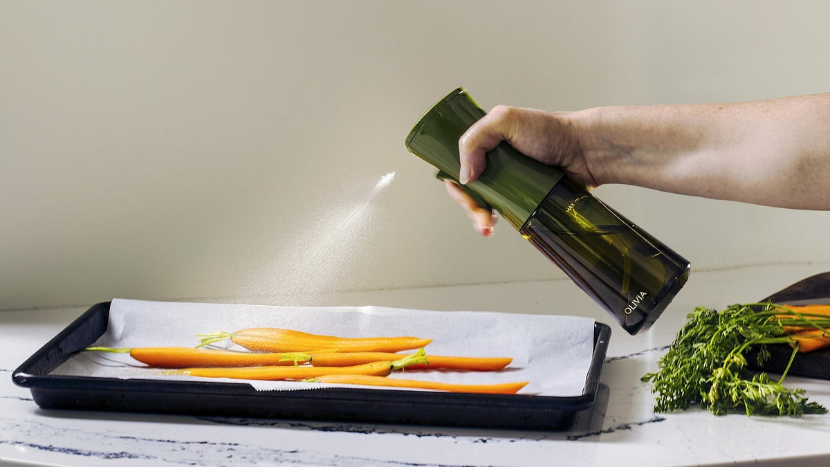 Hand holding a green glass oil sprayer that releases a fine mist over whole carrots on a parchment-lined baking tray