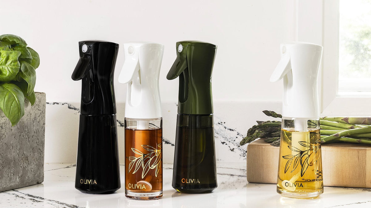 Four Olivia oil sprayers in black, clear, olive green and clear glass lined up on a marble kitchen counter beside basil and asparagus