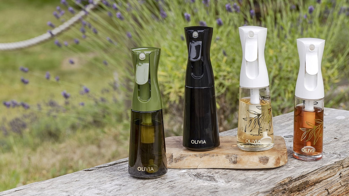 Four glass oil sprayer bottles filled with olive oil and lighter cooking oils on a wooden board outdoors, lavender behind